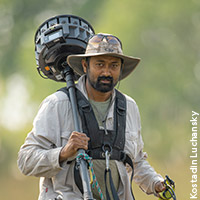 Photo of Sandesh Kadur in the field wearing khaki clothes and a hat, carrying equipment on his shoulder