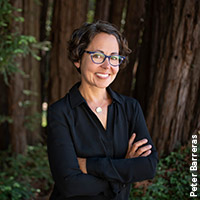 Photo of Beth Shapiro, smiling, arms crossed, wearing a black top and glasses, with trees in the background