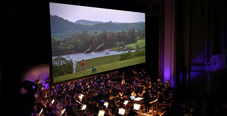 "Jurassic Park" shown on movie screen with symphony playing in front