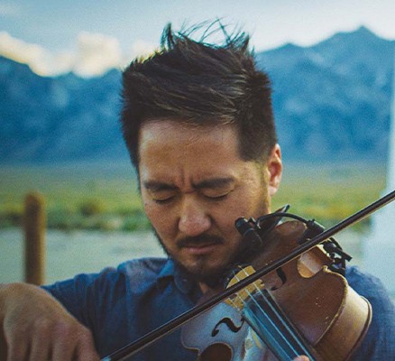 Photo of Kishi Bashi playing violin with a soft focus monument and mountains in the background.