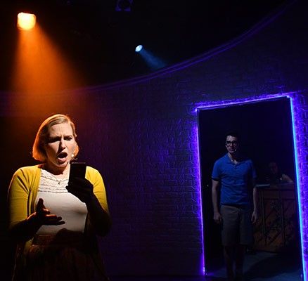 Tinderella production photo of shocked-looking actor holding phone with another actor lurking in a doorway