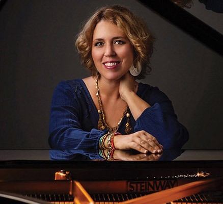 Photo of Gabriela Montero seated at a grand piano against a grey background.
