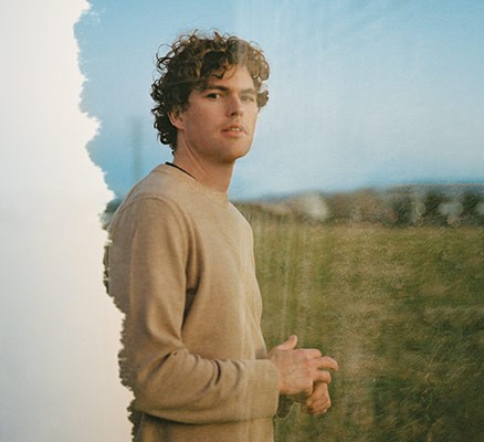 Photo of Vance Joy (white, male, with curly, brown hair) wearing a beige sweater