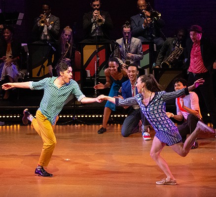 Photo of a male and a female dancer swing dancing together with band in back