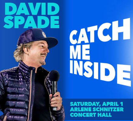 Photo of David Spade holding microphone, wearing puffy coat and trucker hat