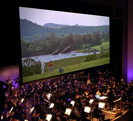 Photo of scene from Jurassic Park shown on screen with symphony performing