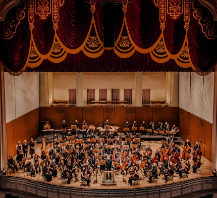 symphony members sit on stage with instruments facing camera