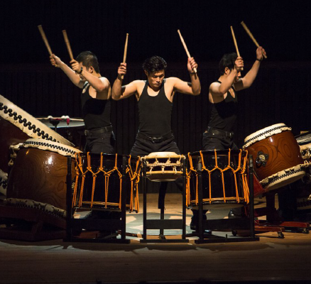 three kodo drummers behind large drums with sticks in air