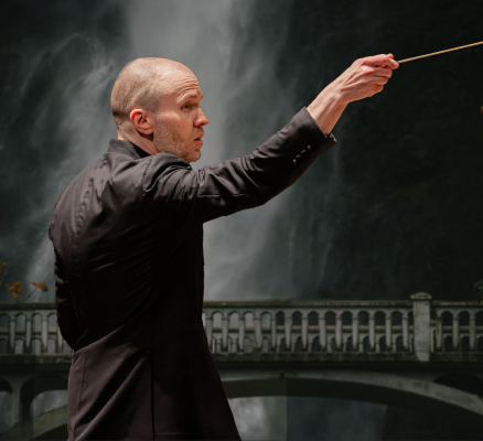 conductor facing right in front of backdrop of darkened photo of a bridge in front of a waterfall