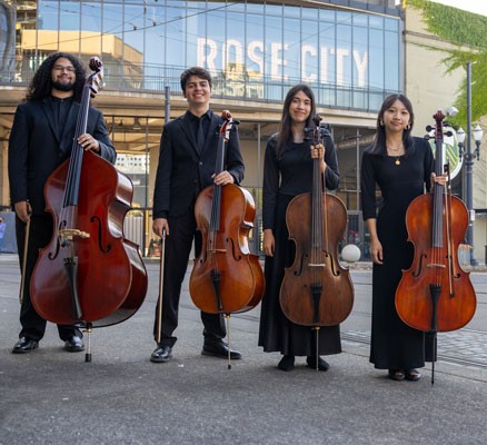 Four youth cello players stand on a street with their instruments