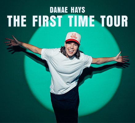 Photo of Danae Hays bowing in a bright spotlight with her arms spread and wearing a white t-shirt and baseball cap.
