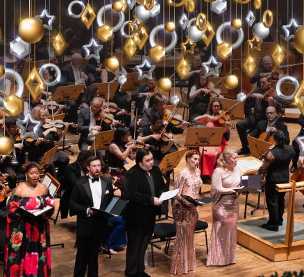 Choir singers perform in front of the Oregon Symphony. Hanging ornaments adorn the image.