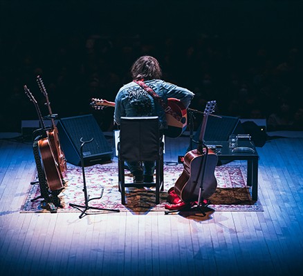 A photo of Ryan Adams on a dark stage with instruments surrounding him. His back is to the camera.