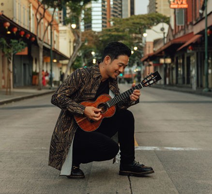 Jake Shimabukuro kneeling in the middle of an empty city street with his ukulele.